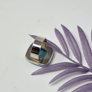 May include: A silver ring with a large, square, light blue stone. The ring band is wide and adjustable. The stone is set in a silver bezel. The ring is displayed on a white surface with a purple leaf.
