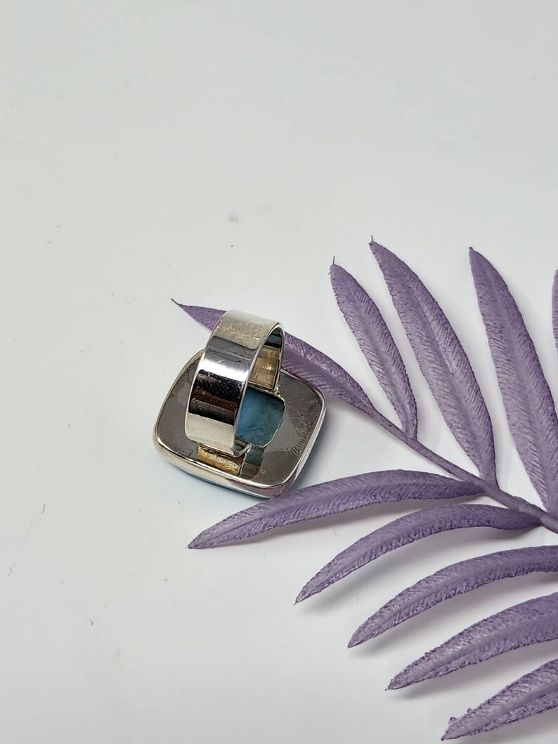 May include: A silver ring with a large, square, light blue stone. The ring band is wide and adjustable. The stone is set in a silver bezel. The ring is displayed on a white surface with a purple leaf.
