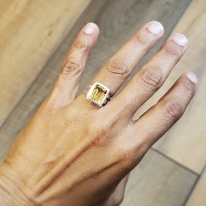 May include: A silver ring with a large, rectangular, yellow gemstone. The ring is set on a hand with a light skin tone, against a wooden background. The gemstone is a vibrant yellow color.