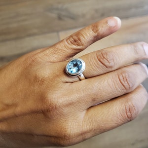 May include: A silver ring featuring a large, oval-cut, light blue gemstone. The gemstone is set in a simple bezel setting, and the ring is displayed on a hand.