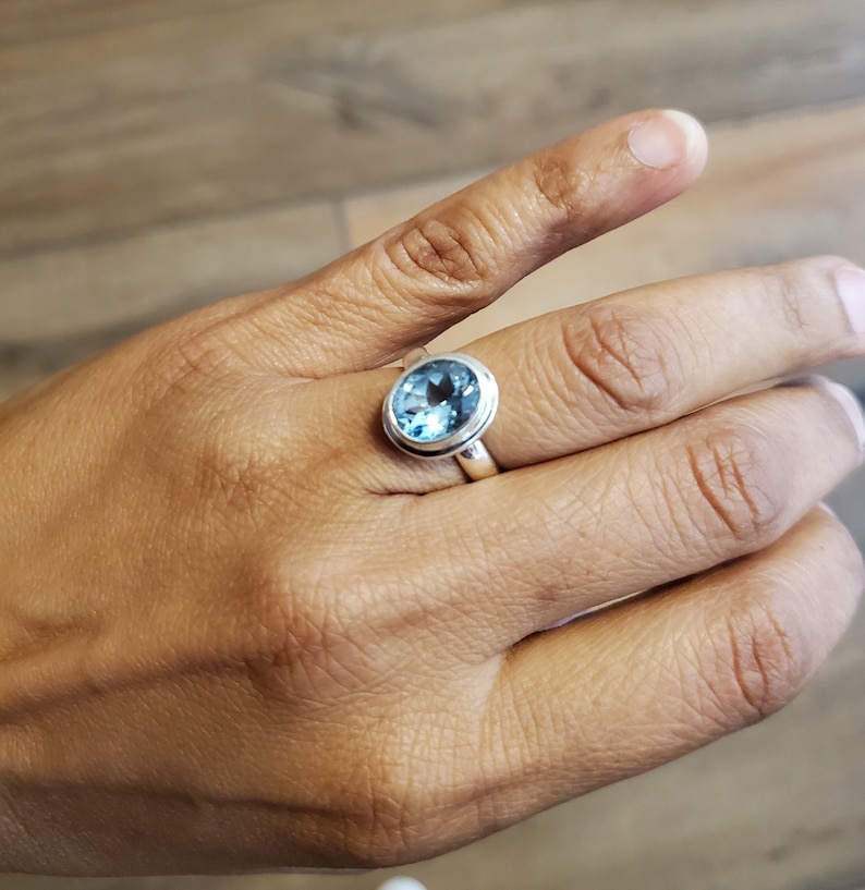 May include: A silver ring featuring a large, oval-cut, light blue gemstone. The gemstone is set in a simple bezel setting, and the ring is displayed on a hand.