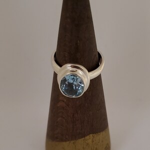 May include: A silver ring with a large, round, light blue gemstone. The gemstone is set in a bezel setting, and the ring is displayed on a brown wooden cone. The ring is a classic design, suitable for everyday wear or special occasions.