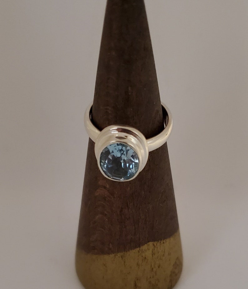 May include: A silver ring with a large, round, light blue gemstone. The gemstone is set in a bezel setting, and the ring is displayed on a brown wooden cone. The ring is a classic design, suitable for everyday wear or special occasions.