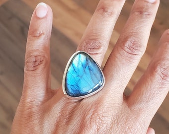 May include: A silver ring featuring a large, triangular labradorite stone. The stone displays iridescent blue and teal hues. The ring's setting is a simple, polished silver band. The ring is shown on a hand.