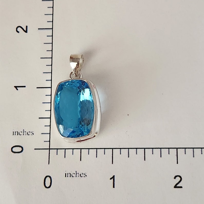 May include: A rectangular, light blue gemstone pendant with a silver-colored metal setting. The pendant is approximately 1 inch wide and 1 inch long. The pendant has a loop at the top for a chain. The gemstone is a bright, clear blue color.