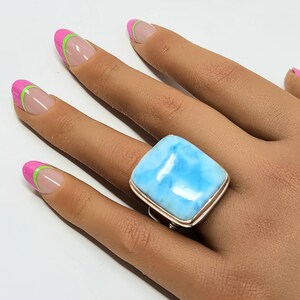 May include: A silver ring with a large, rectangular, light blue gemstone. The ring is set on a hand with manicured nails painted with pink and green accents. The gemstone has a cloudy appearance.