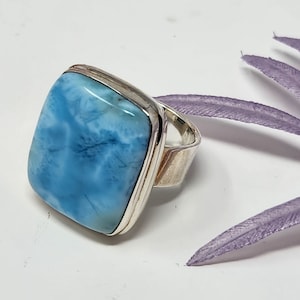 May include: A square-cut, light blue larimar gemstone ring with a silver band. The stone has a marbled appearance with shades of white and blue. The ring is set against a white background with a purple decorative element.