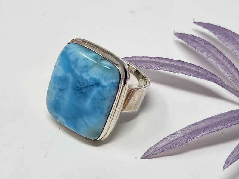 May include: A square-cut, light blue larimar gemstone ring with a silver band. The stone has a marbled appearance with shades of white and blue. The ring is set against a white background with a purple decorative element.
