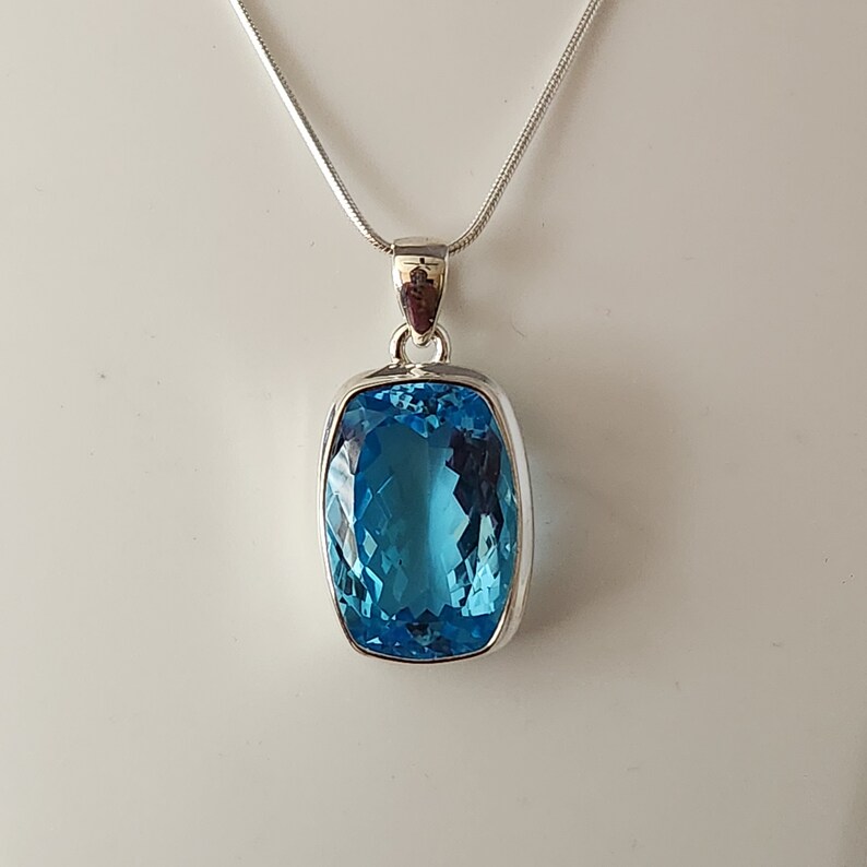 May include: A rectangular, faceted, bright blue gemstone pendant with a silver-colored metal setting hangs from a delicate silver chain. The pendant's facets catch the light, creating a sparkling effect. The jewelry is displayed against a plain white background.