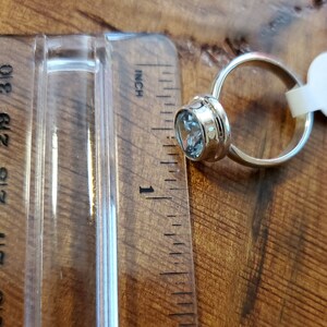 May include: A gold-colored ring with a light blue gemstone, approximately 1 inch in diameter, is displayed next to a ruler. The ring's setting has a decorative design. The background is a wooden surface.