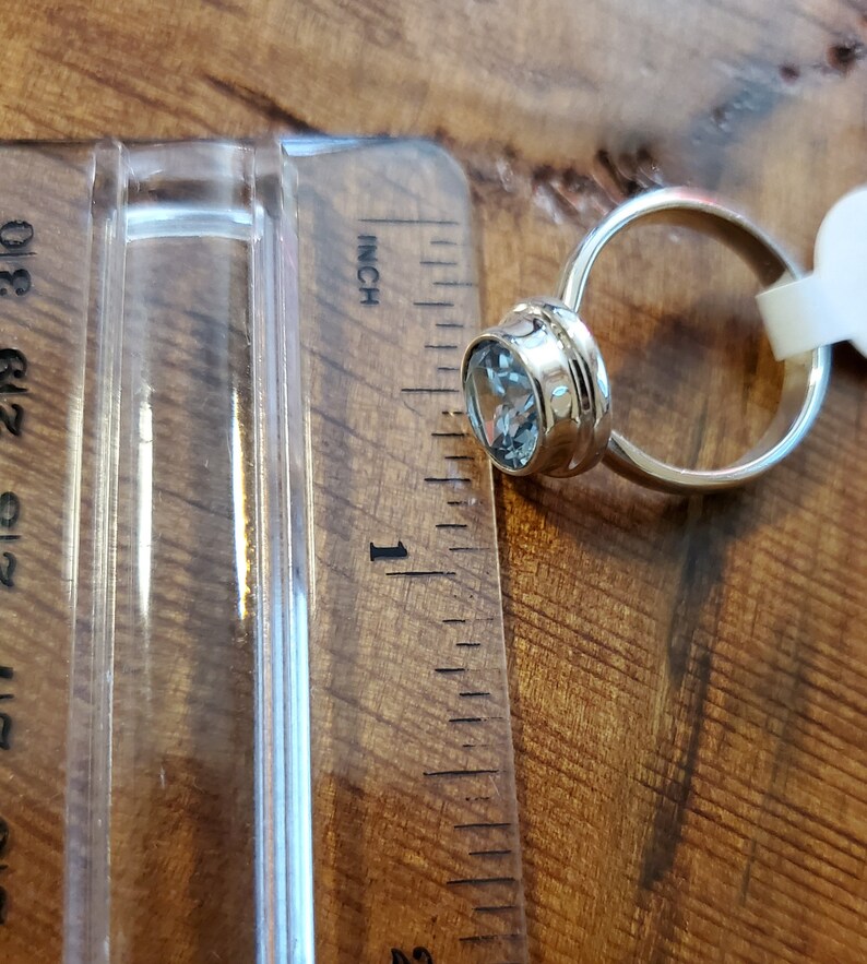 May include: A gold-colored ring with a light blue gemstone, approximately 1 inch in diameter, is displayed next to a ruler. The ring's setting has a decorative design. The background is a wooden surface.