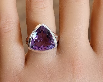 May include: A silver ring featuring a large, triangular amethyst gemstone. The purple gemstone is faceted and set in a polished silver band. The ring is displayed on a hand.