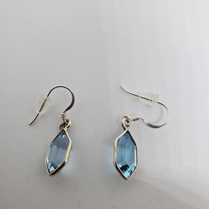 May include: A pair of silver-toned earrings with a light blue, elongated gemstone. The gemstones are set in a silver bezel and hang from a curved ear wire. The earrings are displayed on a white surface.