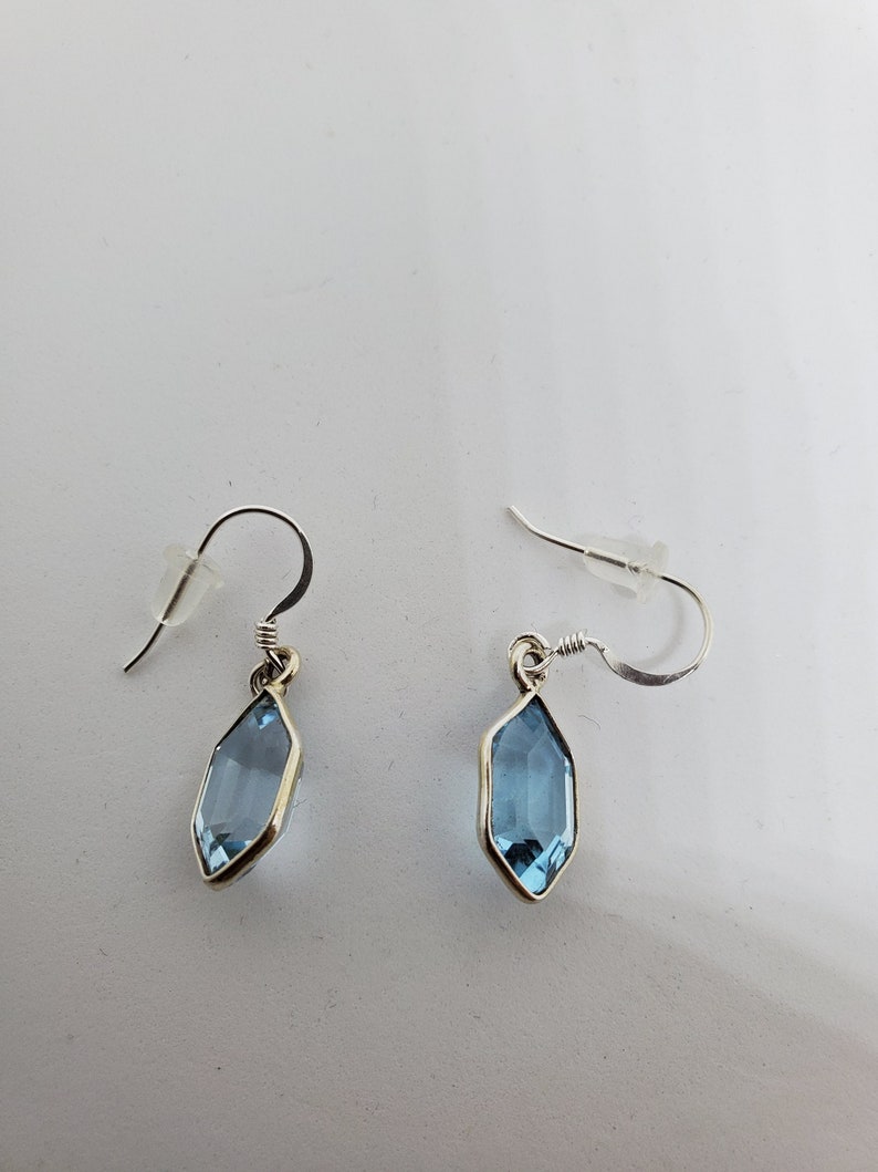 May include: A pair of silver-toned earrings with a light blue, elongated gemstone. The gemstones are set in a silver bezel and hang from a curved ear wire. The earrings are displayed on a white surface.