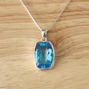 May include: A silver necklace with a rectangular, light blue pendant. The pendant has a faceted surface, and the necklace is displayed on a wooden surface.