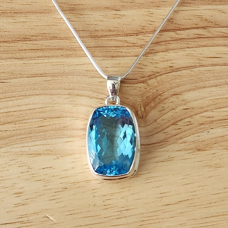 May include: A silver necklace with a rectangular, light blue pendant. The pendant has a faceted surface, and the necklace is displayed on a wooden surface.