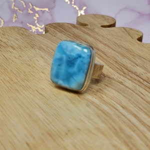May include: A square-cut, light blue larimar gemstone ring with a silver band. The gemstone has a cloudy, ocean-like appearance. The ring is set on a wooden surface.