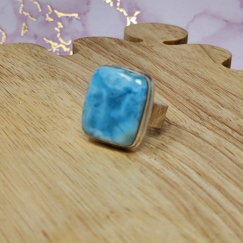 May include: A square-cut, light blue larimar gemstone ring with a silver band. The gemstone has a cloudy, ocean-like appearance. The ring is set on a wooden surface.