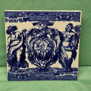May include: A square ceramic tile featuring a blue and white transferware pattern. The design displays a central heraldic crest topped with a crown, flanked by two standing figures. Decorative borders with intricate scrollwork frame the top and bottom.