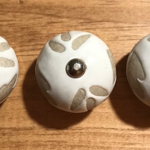 May include: Three white ceramic knobs with a light brown floral design and a silver metal accent in the center.