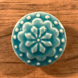 May include: A turquoise blue ceramic knob with a white floral design. The knob has a raised edge and a smooth finish.