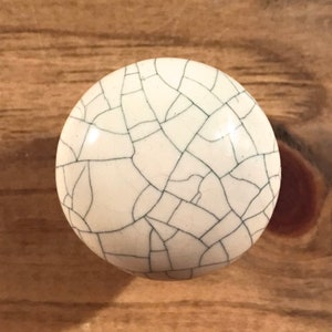 May include: White ceramic knob with a crackle glaze finish. The knob has a circular shape and a white background with a fine network of black cracks.