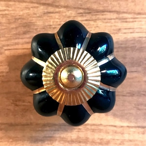 May include: A black ceramic knob with a gold-colored center and a scalloped edge. The knob has a decorative design with gold lines radiating from the center.