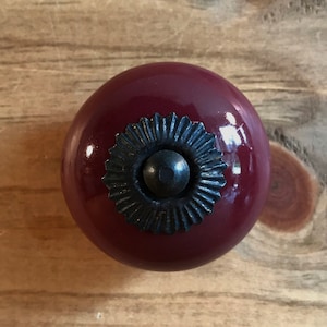 May include: A dark red ceramic cabinet knob with a black metal decorative accent. The knob has a shiny finish.