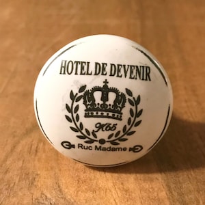 May include: White ceramic knob with black lettering that reads "Hotel de Devenir" with a crown and laurel wreath design. The text "Ruc Madame" is also included.