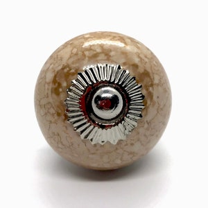 May include: A beige ceramic knob with a silver decorative ring and a silver center. The knob has a speckled pattern.