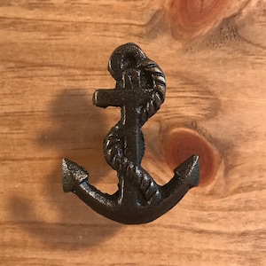 May include: A black metal anchor with a rope design. The anchor is a decorative piece and is mounted on a wooden surface.