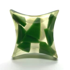 May include: A clear, hourglass-shaped decorative object filled with irregularly shaped, vibrant green pieces. The object has a smooth, curved design and is set against a white background, highlighting the contrast between the clear exterior and the green interior.