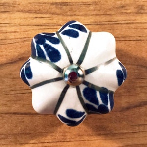 May include: A white and blue floral ceramic cabinet knob. The knob has a flower shape with a silver metal center and a dark blue leaf pattern on the white petals. The knob is set against a wooden background.
