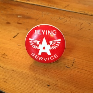 May include: A red and white round button with the text "FLYING A SERVICE" and a white letter "A" with wings on either side.