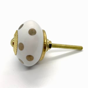 May include: White ceramic cabinet knob with gold-colored hardware and a brass screw. The knob features a pattern of tan polka dots and a decorative gold-colored accent.
