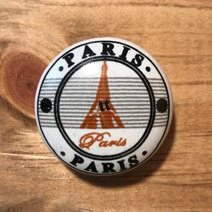 May include: A round, white ceramic knob with a design featuring the Eiffel Tower in orange and the word "Paris" in black. The knob has a striped background and the word "Paris" is repeated around the edge.