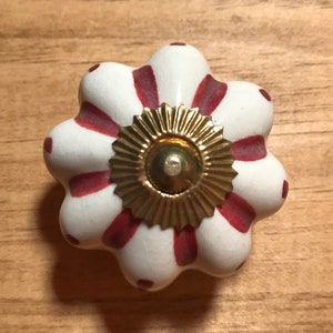 May include: A white and red floral ceramic cabinet knob with a gold-colored metal base. The knob has a flower-like shape with red stripes and dots. The gold base has a sunburst design.