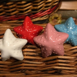 May include: Four ceramic starfish drawer pulls in different colours: white, red, pink, and blue. The starfish are arranged on a woven wicker surface.