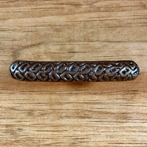 May include: A dark bronze-colored cabinet pull with a textured, woven pattern. The hardware is cylindrical with rounded ends. The pull is set against a light brown wooden surface.