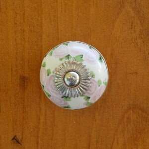 May include: White ceramic knob with a floral design featuring pink roses and green leaves. The knob has a silver metal center.