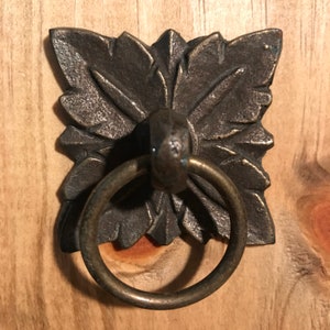 May include: A square, ornate, bronze-coloured metal pull with a ring handle. The pull has a detailed, floral design.