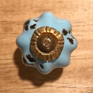 May include: A light blue ceramic knob with a gold-colored metal accent. The knob has a distressed finish with black speckles.