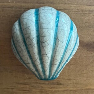 May include: A ceramic seashell-shaped decorative accent with a crackle finish. The shell is white with vertical turquoise stripes. The object is on a wooden surface.