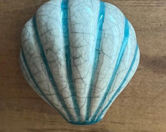 May include: A ceramic seashell-shaped decorative accent with a crackle finish. The shell is white with vertical turquoise stripes. The object is on a wooden surface.