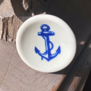 May include: A small white ceramic dish with a blue anchor design. The anchor is a traditional nautical symbol.