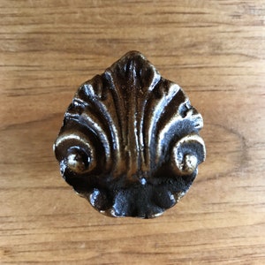 May include: A dark brown metal cabinet knob with a decorative, ornate design. The knob has a scalloped, fan-shaped design with intricate details.
