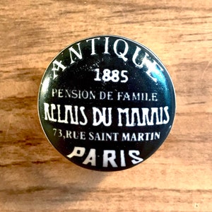 May include: A round, black button with white text. The text reads "ANTIQUE 1885 PENSION DE FAMILLE RELAIS DU MARAIS 73, RUE SAINT MARTIN PARIS". The button is set against a wooden surface.