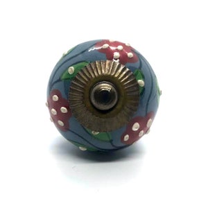 May include: A decorative cabinet knob with a round, blue-gray ceramic body. The knob features a floral design with red flowers, green leaves, and white accents. The base is a bronze-colored metal.