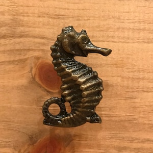May include: A gold-toned metal seahorse figurine with a detailed, textured design. The seahorse is standing upright with its tail curled around its body.