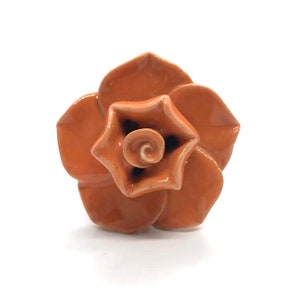 May include: A ceramic flower-shaped decorative object. The flower has a layered petal design in a warm orange hue, with a darker center. The center features a spiral detail. The object is set against a white background.